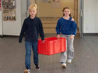 Zwei Kinder tragen gemeinsam eine rote Kiste über den Flur
